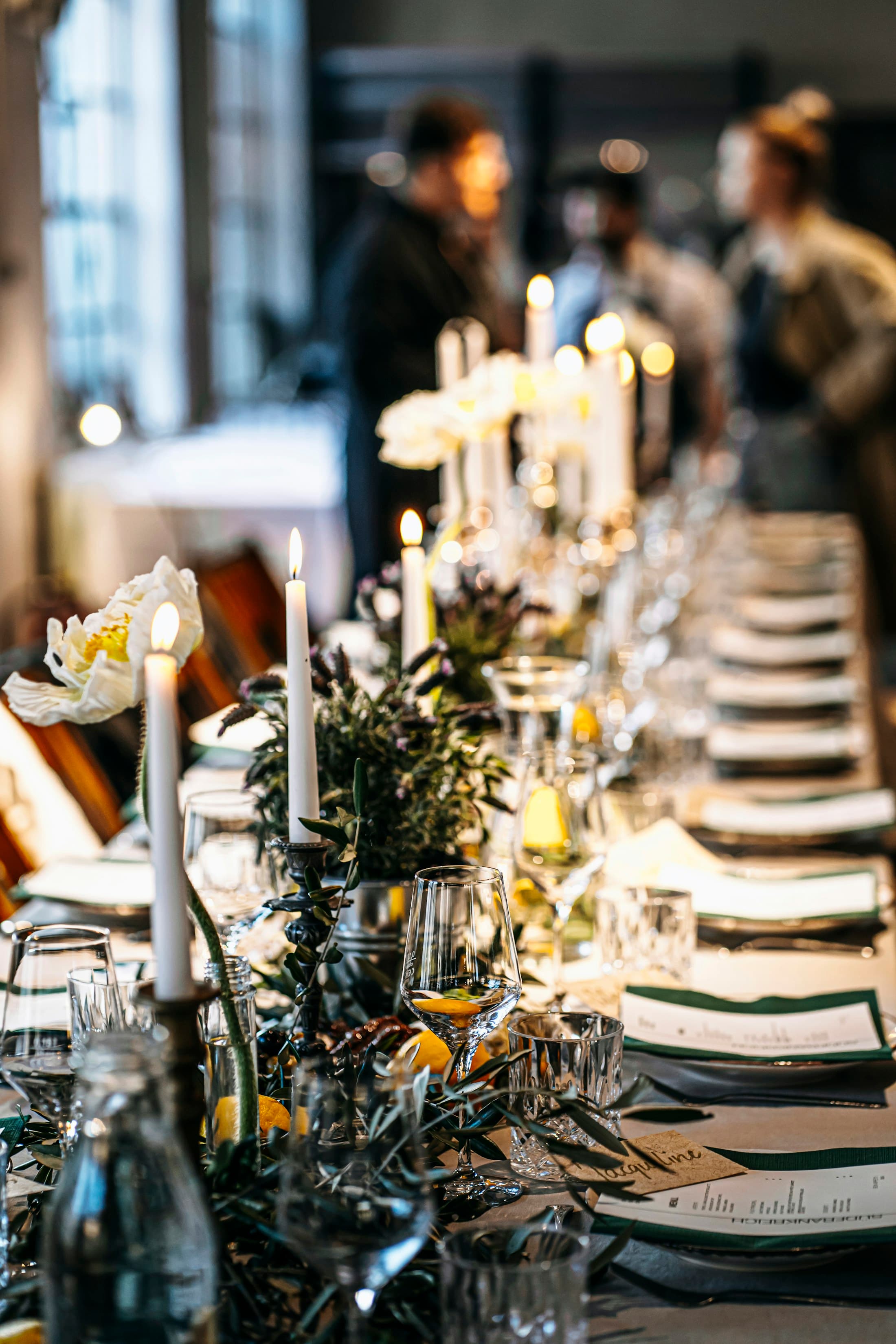 A warm dining table set with taper candles, glassware, flowers, and layered place settings.
